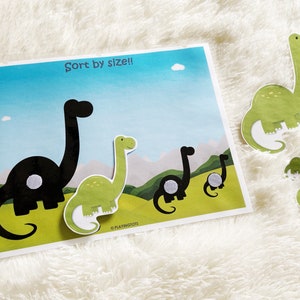 Puede incluir: Un conjunto de cinco piezas de rompecabezas con forma de dinosaurio, cuatro verdes y una negra, con el texto "&iexcl;Ordenar por tama&ntilde;o!" sobre un fondo azul. Las piezas est&aacute;n dise&ntilde;adas para ser ordenadas por tama&ntilde;o, con la pieza m&aacute;s grande en la parte superior y la m&aacute;s peque&ntilde;a en la parte inferior.