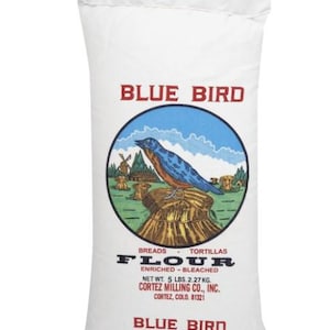 May include: White flour bag with the words "BLUE BIRD" in red and blue. A circular graphic features a bluebird perched on a haystack with a rural landscape background. The bag contains 5 lbs of enriched flour.