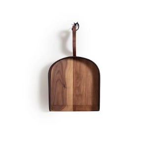 May include: A wooden pizza peel with a dark brown leather handle. The peel has a curved, shovel-like shape with a light and dark wood grain pattern. The handle is attached to the top of the peel with a metal ring.