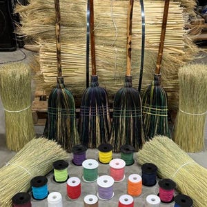 May include: Four handcrafted brooms with long wooden handles and bristles in shades of black, green, and yellow, arranged in front of a large bundle of straw. Below are numerous spools of colorful thread in various hues including red, blue, and white.