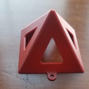 May include: A red, triangular-shaped ornament with a cut-out design. The ornament has a small loop at the bottom for hanging.