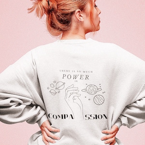 May include: A white sweatshirt with a graphic print of a hand holding planets and the text "There is so much power in compassion".