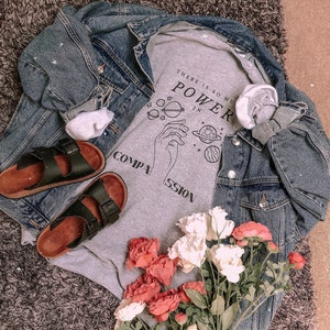 May include: Gray t-shirt with a graphic of a hand holding a planet with the text "There is so much power in compassion". The t-shirt is layered over a denim jacket and paired with black sandals and a bouquet of pink and white flowers.