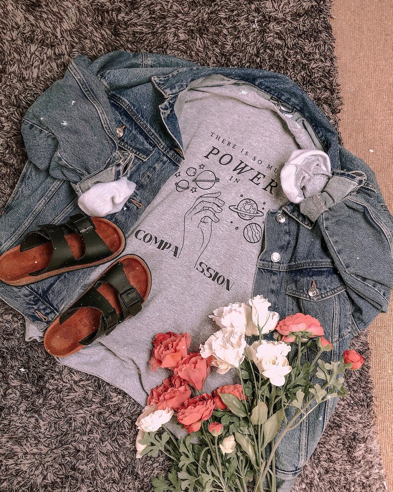 May include: Gray t-shirt with a graphic of a hand holding a planet with the text "There is so much power in compassion". The t-shirt is layered over a denim jacket and paired with black sandals and a bouquet of pink and white flowers.