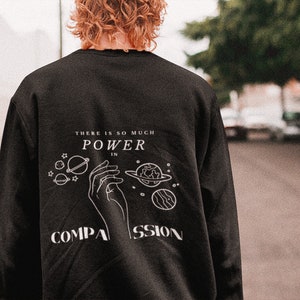 May include: Black sweatshirt with a white graphic design featuring a hand holding planets and stars. The text reads "There is so much power in compassion."