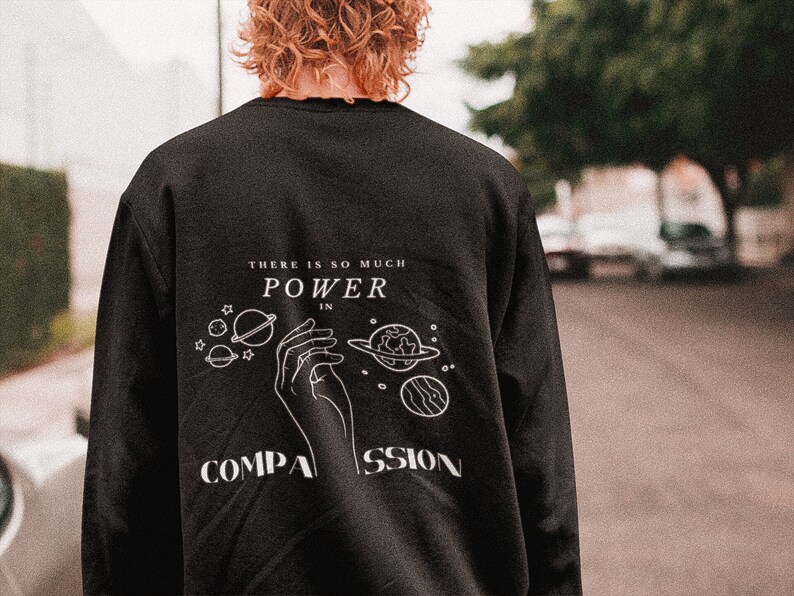 May include: Black sweatshirt with a white graphic design featuring a hand holding planets and stars. The text reads "There is so much power in compassion."