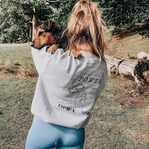 May include: A woman wearing a grey sweatshirt with the text "There is so much power in compassion" and a black and white graphic of a hand with planets around it. She is holding a brown and black dog in her arms.