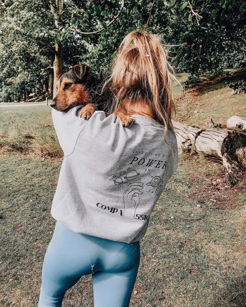 May include: A woman wearing a grey sweatshirt with the text "There is so much power in compassion" and a black and white graphic of a hand with planets around it. She is holding a brown and black dog in her arms.