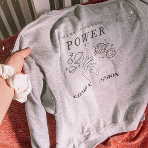 May include: Gray sweatshirt with a graphic print of a hand holding planets and the text "There is so much power in compassion".