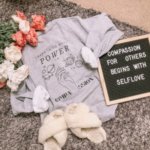 May include: Gray sweatshirt with a graphic of hands holding planets and the text "There is so much power in compassion". A black letter board with the text "Compassion for others begins with self love" is in the background.