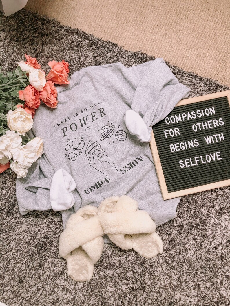 May include: Gray sweatshirt with a graphic of hands holding planets and the text "There is so much power in compassion". A black letter board with the text "Compassion for others begins with self love" is in the background.