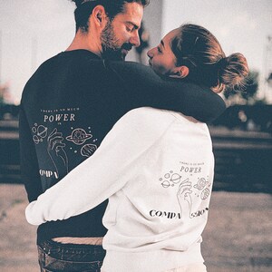 May include: A couple wearing matching sweatshirts with a graphic of a hand holding planets and the text "There is so much power in compassion".