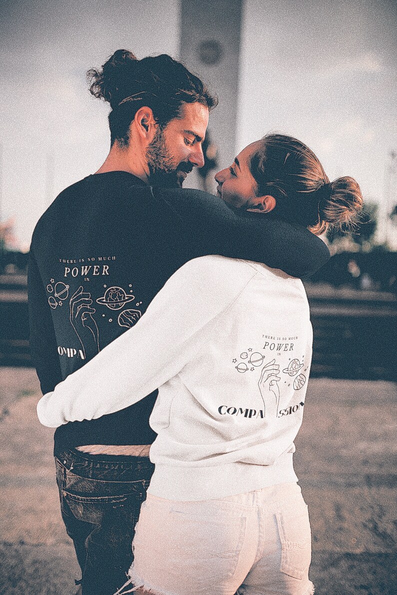 May include: A couple wearing matching sweatshirts with a graphic of a hand holding planets and the text "There is so much power in compassion".