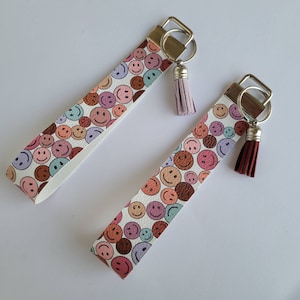 May include: Two colorful keychains with a white background and a pattern of smiling faces. The keychains have silver key rings and tassels. One tassel is pink and the other is burgundy.
