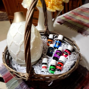 Pode incluir: Uma cesta de vime cheia de garrafas de óleo essencial em várias cores, incluindo verde, azul, vermelho e roxo. A cesta está sentada em uma mesa com um difusor branco e um vaso de flores.