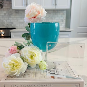 May include: A teal glass vase, a clear acrylic frame, and a book titled "Beautifully Organized" are arranged with faux flowers. The flowers include white, pink, and peach peonies and ranunculus. The scene is set on a white countertop.