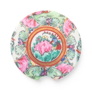 May include: A round, white ceramic coaster with a colorful floral design. The coaster features pink roses, green leaves, and a central medallion with a pink flower and a bird. The design is framed by a gold border.