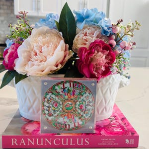May include: A white vase filled with pink, light blue, and white flowers. A decorative plate with a colourful design rests on a pink book titled "RANUNCULUS". The arrangement is set on a white countertop.