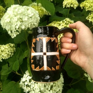 May include: A glossy black ceramic mug featuring a central geometric design composed of tan and white triangular and cross-shaped patterns. The mug has a curved handle on the right side and a smooth, reflective finish throughout its exterior surface.