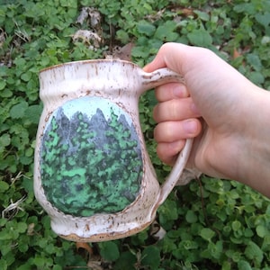 May include: A handmade ceramic mug with a forest scene painted on the front. The mug is off-white with brown accents and a large handle. The forest design features green trees and a blue sky.