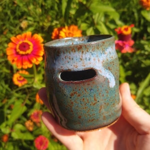 May include: A small, cylindrical ceramic vessel with a mottled teal and brown glaze, featuring a horizontal rectangular cutout near the top rim. The surface has a glossy finish with subtle blue highlights and a speckled texture throughout the body.