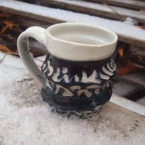 May include: A white ceramic mug with a black and white patterned design. The mug has a rounded body and a handle.