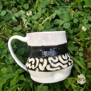 May include: A white ceramic mug with a black band and white swirl design. The mug has a handle and is sitting on a bed of green clover.