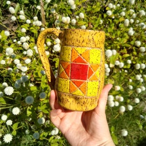 May include: A yellow ceramic mug with a speckled finish and a handle. The front features a geometric star pattern in shades of red, orange, and yellow, composed of square and triangular segments with a speckled texture throughout the design.