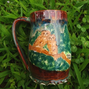 May include: A brown ceramic mug with a green, brown, and white design of two owls perched on a branch. The mug has a brown glaze and a scalloped rim.