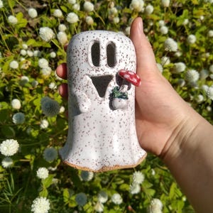 May include: A white ceramic ghost figurine with brown speckles, featuring cut-out eyes and a triangular mouth. The ghost has a small, sculpted red and white mushroom attached to its side, with a brown clay base visible at the bottom edge.