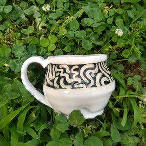 May include: White ceramic mug with a black and white abstract design. The mug has a rounded body and a handle.