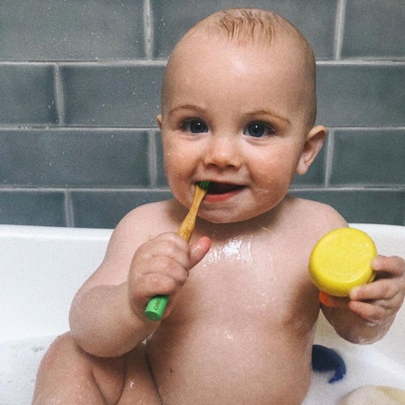 May include: A baby in a bathtub is brushing their teeth with a green and wooden toothbrush. The baby is smiling and has a yellow rubber ducky toy in their other hand.