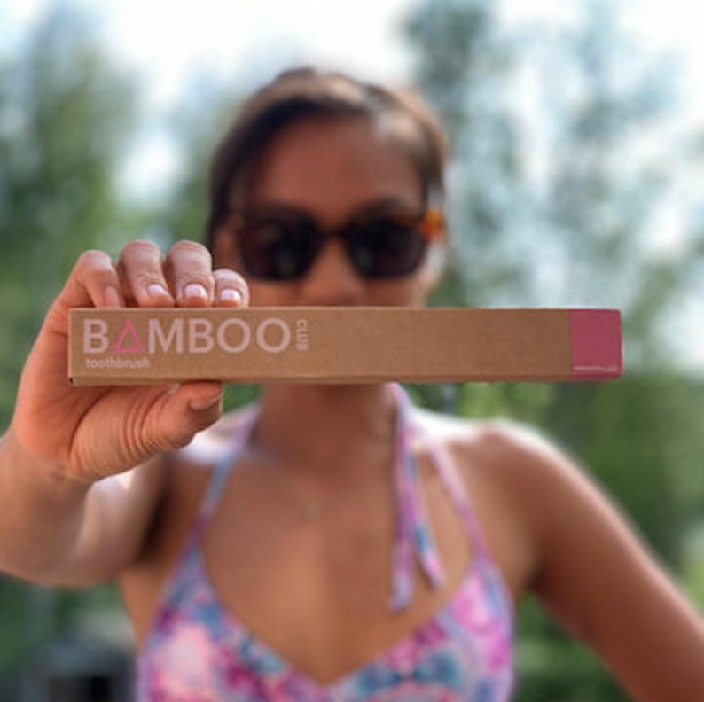 May include: A person holding a brown cardboard box with the text "BAMBOO club toothbrush" printed on it in white and pink.
