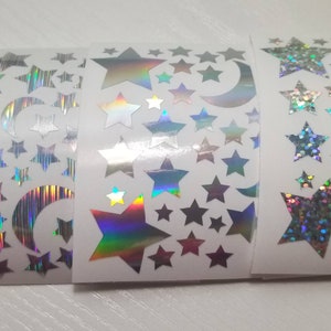 以下が含まれることがあります： 星と三日月が描かれた虹色のホログラフィックステッカーが3ロール。ステッカーは白い背景です。