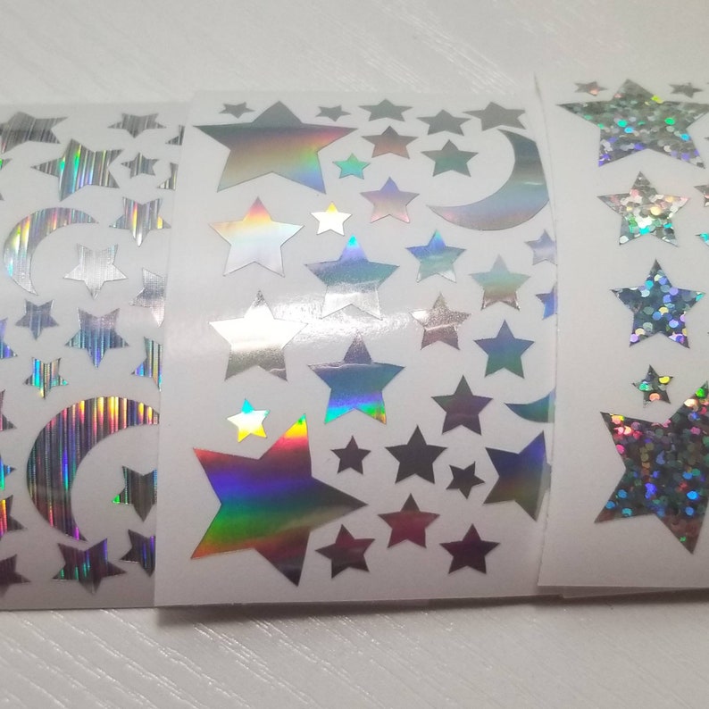 以下が含まれることがあります： 星と三日月が描かれた虹色のホログラフィックステッカーが3ロール。ステッカーは白い背景です。