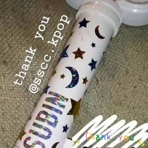 以下が含まれることがあります： 白いプラスチック製のチューブに、ホログラフィックステッカーが貼られています。ステッカーには「SUBIN」と書いてあり、金色の星と銀色の月のデザインが施されています。チューブには「thank you @sscc.kpop」という文字が書かれています。