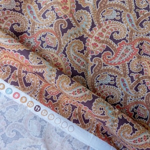 May include: A close-up of a brown paisley patterned fabric with a white border. The fabric is folded over and the border has numbers printed on it.
