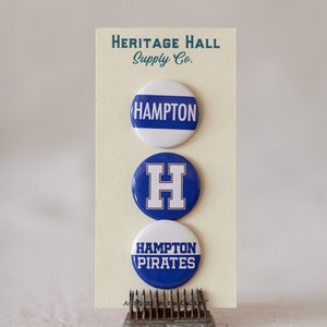 May include: Three blue and white button pins with the text "Hampton" and "Hampton Pirates" and a blue "H" on a white background. The pins are on a white card with the text "Heritage Hall Supply Co."