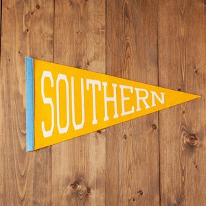 May include: A yellow felt pennant with the word "SOUTHERN" in white block letters. The pennant has a light blue border and is hanging against a wooden background.