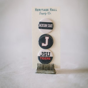 May include: Three round buttons with the text "JACKSON STATE", a letter "J", and "JSU TIGERS" on a cream-colored card. The buttons are black and white, with red accents. The card is held by a small, metal floral frog.