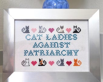 Puede incluir: Un bordado de punto de cruz enmarcado con el texto "CAT LADIES AGAINST PATRIARCHY" en color turquesa. El diseño incluye siluetas de gatos y corazones rosas. El marco es plateado y el fondo es blanco.