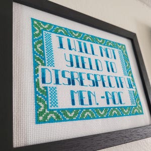 Puede incluir: Obra de arte en punto de cruz enmarcada con el texto "I WILL NOT YIELD TO DISRESPECTFUL MEN - AOC" en azul y verde sobre un fondo blanco. La obra est&aacute; enmarcada en un marco negro.