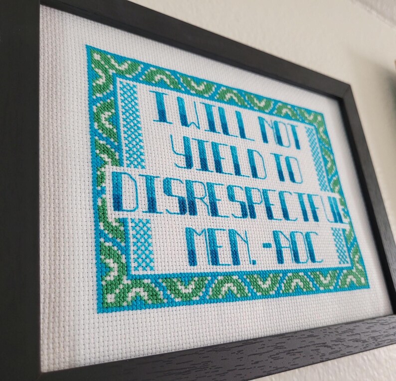 Puede incluir: Obra de arte en punto de cruz enmarcada con el texto "I WILL NOT YIELD TO DISRESPECTFUL MEN - AOC" en azul y verde sobre un fondo blanco. La obra est&aacute; enmarcada en un marco negro.