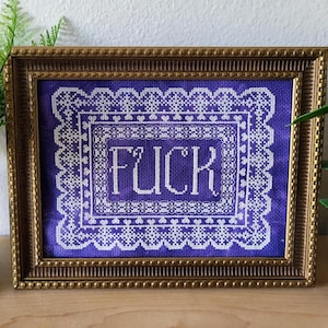 Peut inclure: Une œuvre d'art encadrée au point de croix avec le mot "FUCK" en blanc sur un fond violet. Le motif est encadré d'un cadre doré orné. La broderie au point de croix a une bordure décorative.