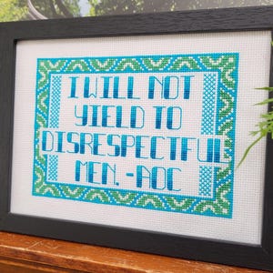 Puede incluir: Obra de arte de punto de cruz enmarcada. El texto "I WILL NOT YIELD TO DISRESPECTFUL MEN. - AOC" est&aacute; bordado en azul y turquesa sobre un fondo blanco. El marco negro es rectangular y de madera.