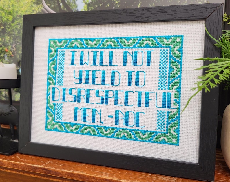 Puede incluir: Obra de arte de punto de cruz enmarcada. El texto "I WILL NOT YIELD TO DISRESPECTFUL MEN. - AOC" est&aacute; bordado en azul y turquesa sobre un fondo blanco. El marco negro es rectangular y de madera.