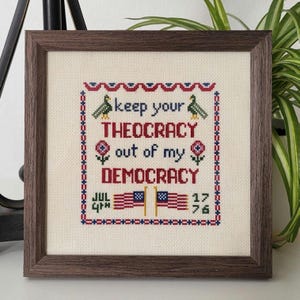 Pode incluir: Um bordado de ponto cruz emoldurado com fundo branco e bordado em vermelho, azul e verde. O texto diz "Keep your theocracy out of my democracy." O bordado também inclui dois pássaros estilizados, flores e a data "Jul 4th 1776".