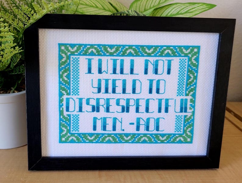 Puede incluir: Arte de punto de cruz enmarcado con la cita "I WILL NOT YIELD TO DISRESPECTFUL MEN. -AOC" en azul y verde. El texto est&aacute; centrado sobre un fondo blanco, enmarcado en negro. El dise&ntilde;o presenta un borde decorativo.