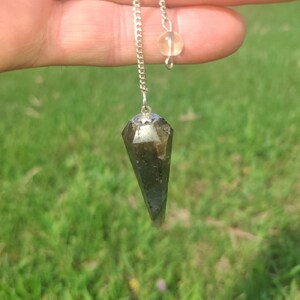 May include: A faceted, dark green crystal pendulum suspended from a silver chain. A small, round, yellow bead is attached to the chain above the crystal. The pendulum is held against a background of green grass.
