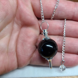 May include: Black agate pendulum with a silver chain and a clear crystal at the end. The pendulum is held in a hand.